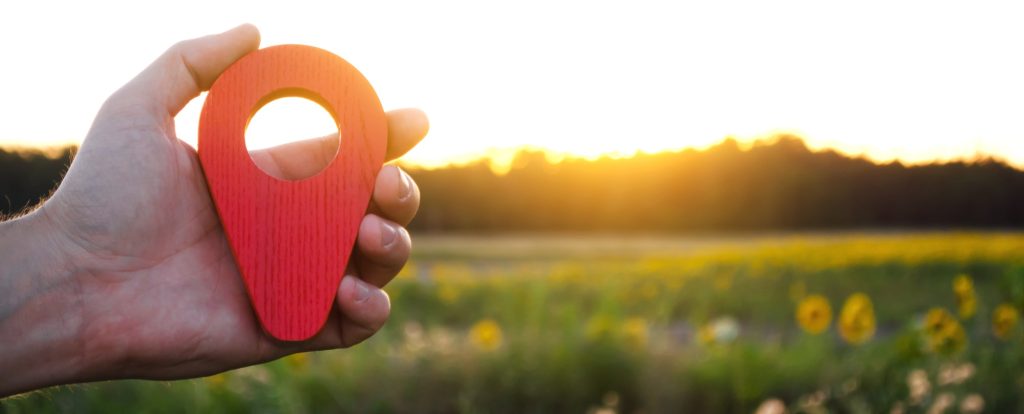 A hand is holding a red location marker in the sunset background